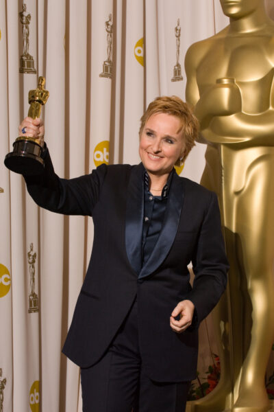 Melissa Etheridge in a tuxedo holding an Oscar with a large Oscar standing behind her.